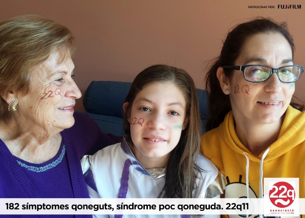 Peu de foto, logo de l’Associació Catalana de la Síndrome 22q/11. Fotografía avia, nena i mare amb el nom de la síndrome escrit a les seves galtes. 